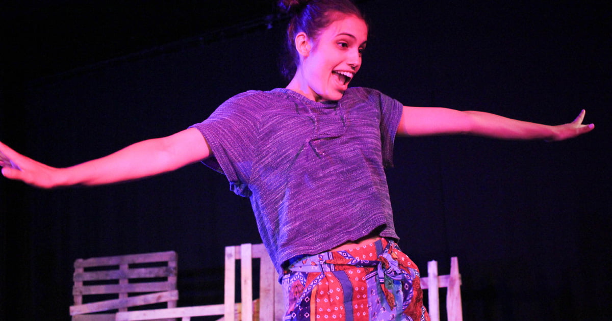 Alma Crespo as Sonia in Ensemble in Godspell (Musical, Edinburgh Fringe, 2017) - Dancing girl with arms stretched wide, wearing a grey t-shirt and multi-coloured patchwork trousers