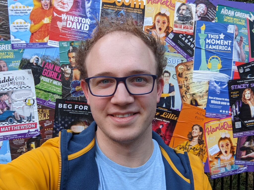 Jack Dryden - Fringe Theatre Producer stood in front of an Underbelly poster wall at the Edinburgh Fringe